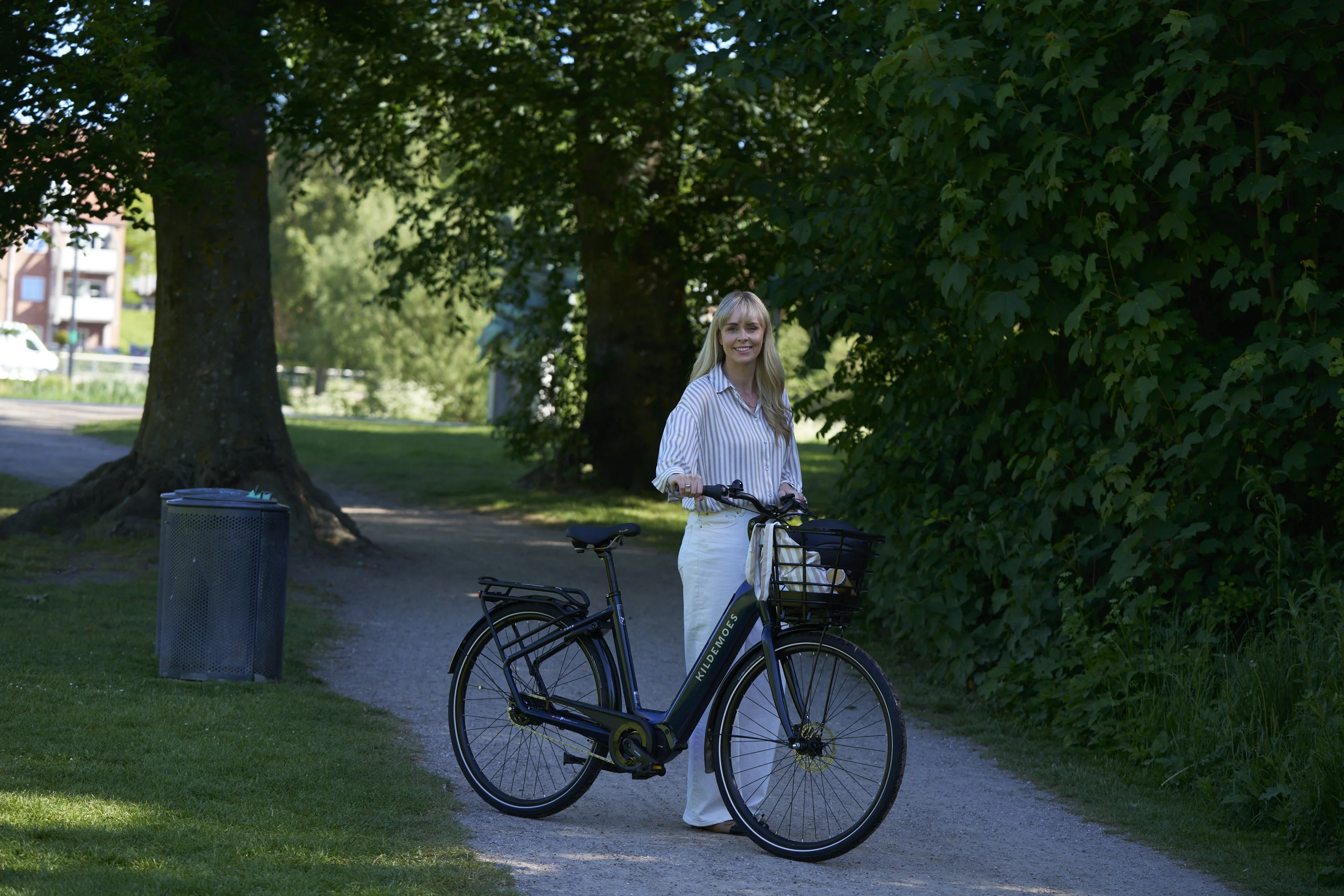 Kvinde står ved en sort Kildemoes damecykel med kurv på en parksti